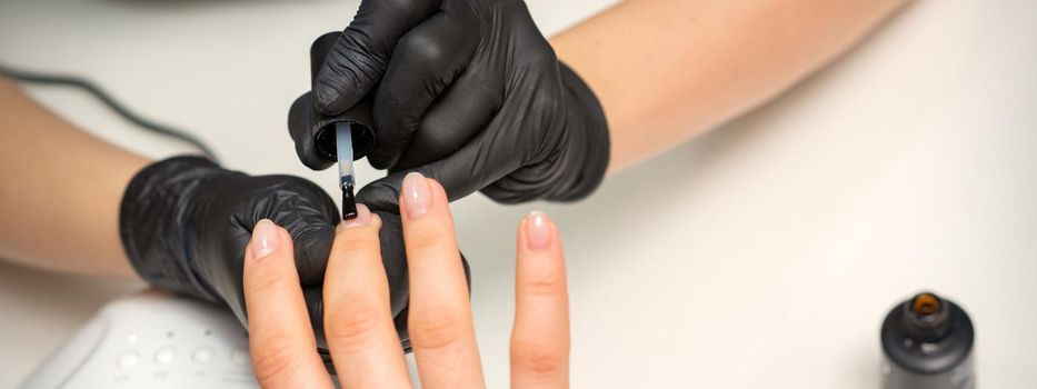 Painting nails of a woman. Hands of Manicurist in black gloves applying transparent nail polish on female Nails in a beauty salon