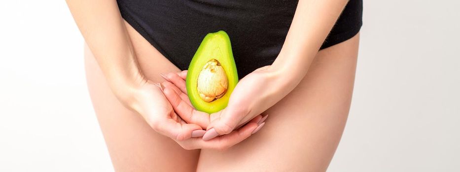 Young woman holding one half of a fresh avocado fruit close to her belly against a white background, healthy nutrition and pregnancy concept