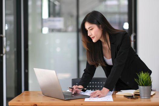 Happy young Asian businesswoman standing using laptop computer at office..