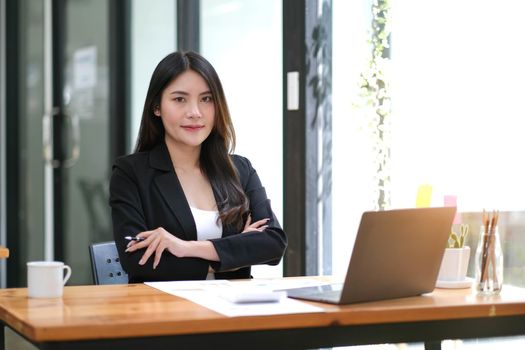 Confident young Asian business woman sitting with arms crossed smiling looking at camera in the office..