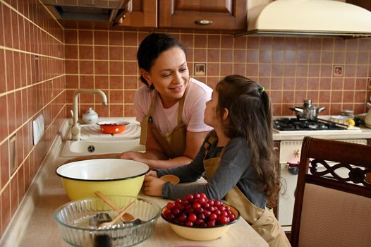 Multi-ethnic cute woman, loving mom talking with her adorable little daughter, standing together by a kitchen countertop, with fresh ingredients for making homemade cherry pie. Child learning cooking