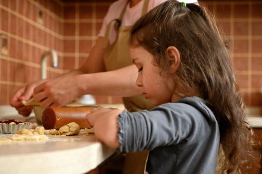 Beautiful European little girl with long hair, using rolling pin, rolling out the dough, cooking a delicious homemade cherry pie with her mother, standing by a kitchen countertop in the home interior