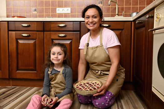 Lovely charming beautiful tender gentle mom and her adorable kid, a little daughter holding in hands pies with sweet cherry berries, smiling at camera, sitting together on the floor of home kitchen