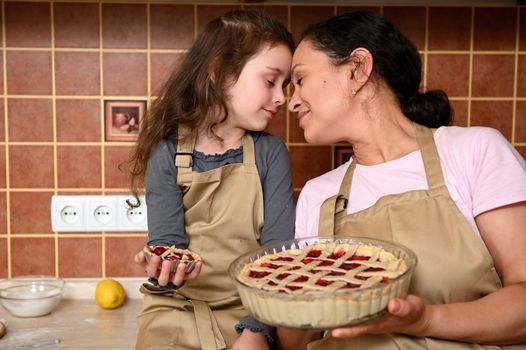 Loving mother and her cute little daughter present their homemade baked pie from cherries to camera, standing close to each other in the home kitchen. Baby girl prepared her first cherry berry cake