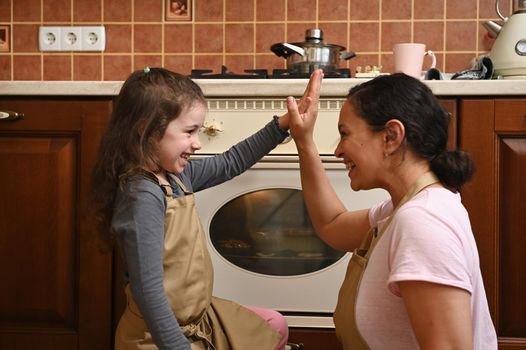 Beautiful young Hispanic woman, a loving mom and her cute little daughter, both of them wearing beige chef aprons, high five while standing by the oven in home kitchen, baking homemade cherry pie