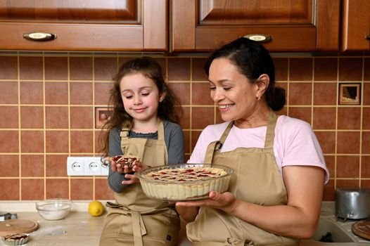 Beautiful young dark-haired multi-ethnic woman, a loving mother smiling cheerfully, standing near her cute little girl holding her first baked little cherry pie, sitting on the kitchen countertop