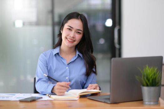 smiling beautiful young Asian businesswoman sitting with laptop and computer while doing some paperwork at the office.