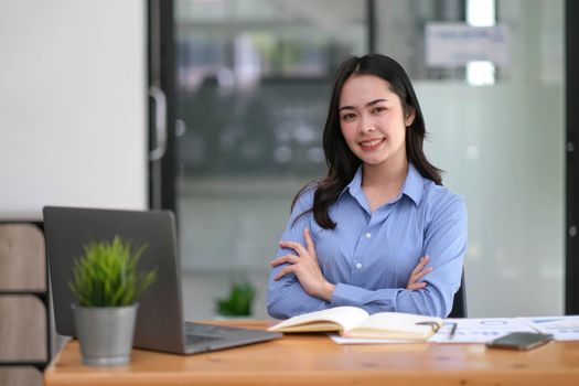 Confident young Asian business woman sitting with arms crossed smiling looking at camera in the office..