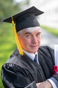 Portrait of an elderly man in a graduation gown and with a diploma in his hands outdoors. Vertical