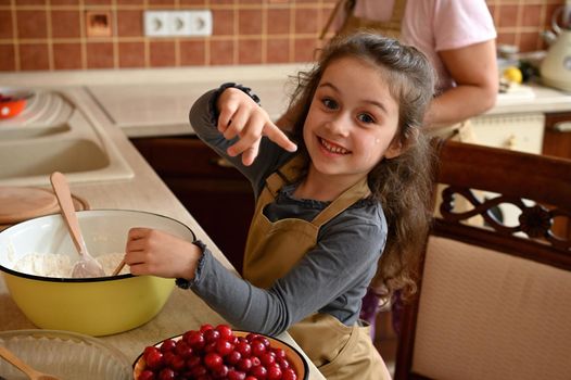 Adorable Caucasian little girl in a beige chef's apron, standing on a chair by a kitchen counterrop with fresh ingredients and smiling a beautiful toothy smile while kneading dough in the home kitchen