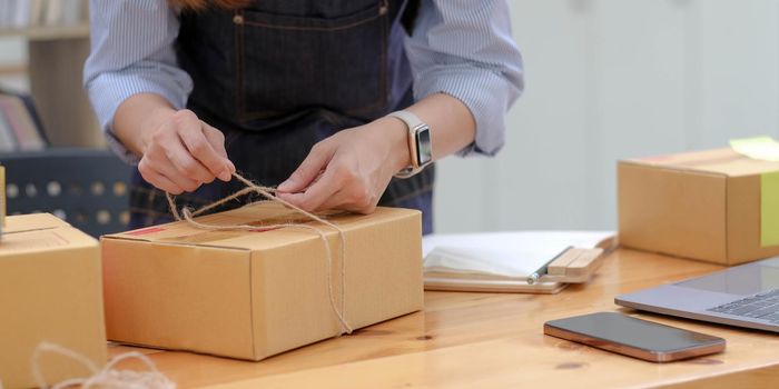 Small startup business concept. Woman tying ropes on a parcel box packs items in preparation for shipping an order. Preparing to ship for e-commerce sellers..
