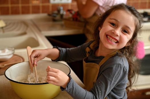 Portrait of a Caucasian cheerful and mischievous little confectioner, an adorable baby girl in a chef's apron, smiling cheerfully, looking at the camera and kneading dough to bake a festive cherry pie