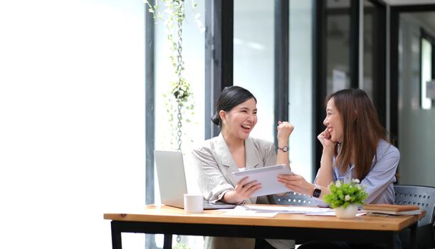 Two young Asian businesswomen show joyful expression of success at work smiling happily with a laptop computer in a modern office..