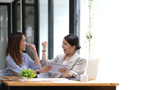 Two young Asian businesswomen show joyful expression of success at work smiling happily with a laptop computer in a modern office..