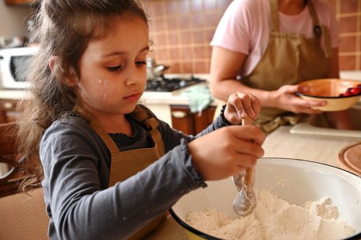Close-up portrait of an adorable Caucasian little girl mixing dry ingredients with white flour in an enamel bowl, kneading dough in home kitchen, helping her mother in cooking festive tasty cherry pie