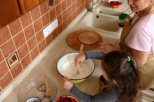 Overhead view: charming little girl standing by countertop, kneading dough for delicious festive cherry pie, helping her mother in the kitchen. Child learns culinary. Mom and daughter cooking together