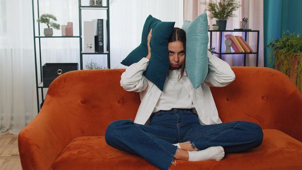 Repair work at neighbours concept. Irritated woman sit on couch cover ears with pillows annoyed by noisy neighbors suffer from headache wishes silence. Thin walls at home flat without sound insulation