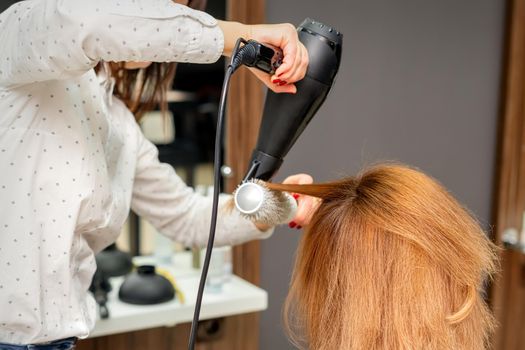Drying hair in the hair studio. Female hairdresser stylist dries hair with a hairdryer and round brush red hair of a woman in a beauty salon