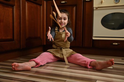 Selective focus on charming child, beautiful little girl, wearing beige chef apron, sitting barefoot on the floor by the oven, in the home rustic kitchen and throwing spoons smiling looking at camera