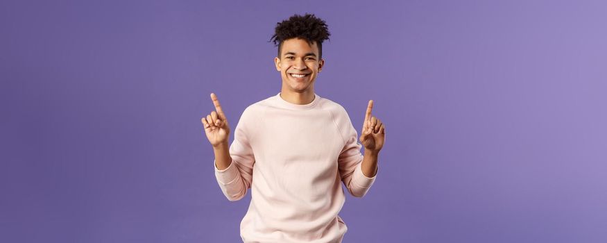 Waist-up portrait of joyful good-looking hispanic man with dreads, promoting product or company banner hanging on top, pointing fingers up, smiling satisfied, recommend subscribe or click link.