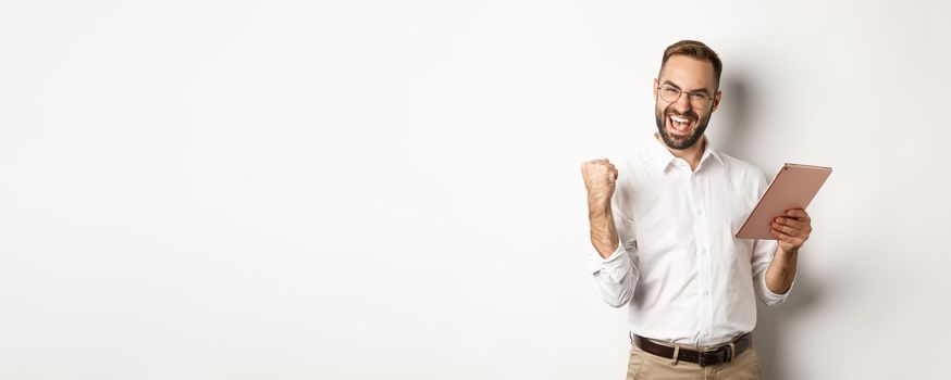 Successful businessman rejoicing on winning online, reading on digital tablet and making fist pump, triumphing, standing over white background.