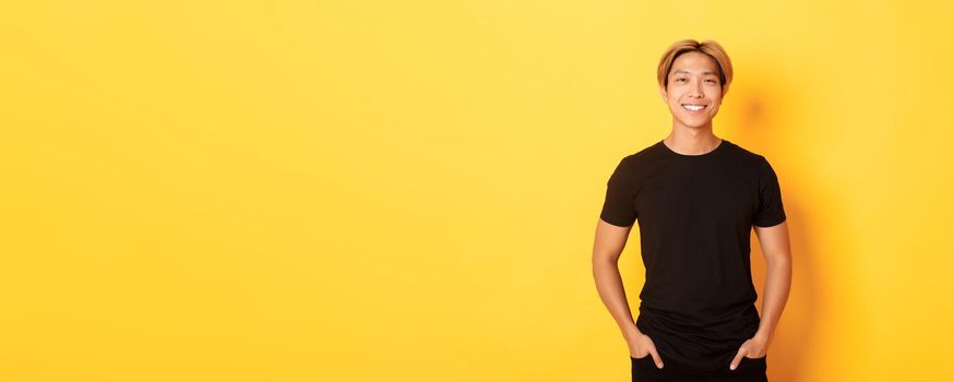 Attractive smiling asian man in black t-shirt, standing pleased, yellow background.