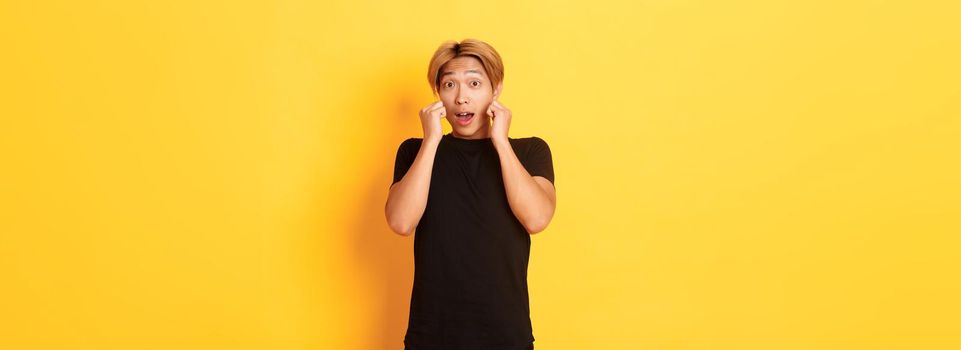 Excited handsome blond guy in black clothes, looking tempted and standing over yellow background.