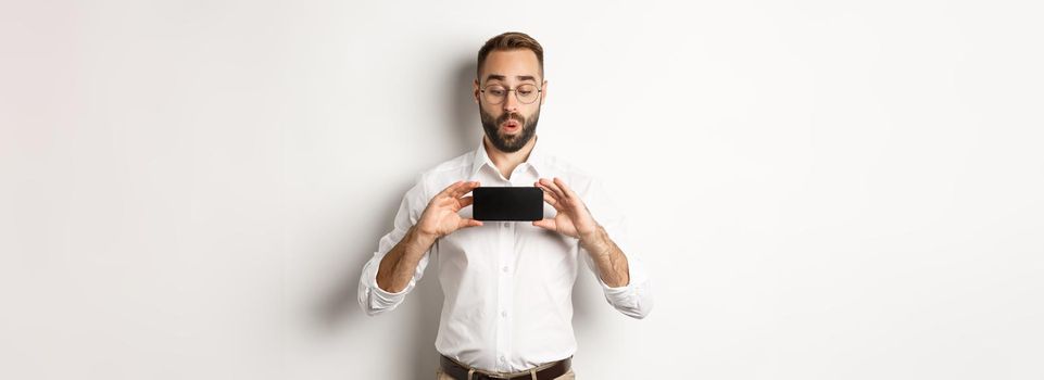 Amused handsome guy showing mobile screen, looking excited at online website, standing over white background.