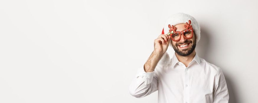 Close-up of handsome bearded guy in xmas party glasses and santa hat, smiling and wishing merry christmas, standing over white background.
