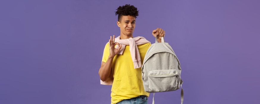 Good but not the best. Portrait of disappointed, unsatisfied young teenage student, guy grimacing without enthusiasm show okay, received new ugly backpack, standing purple background.