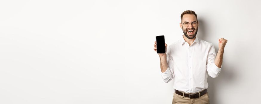 Successful business man showing mobile screen, rejoicing on winning online prize, achieve app goal, standing against white background.