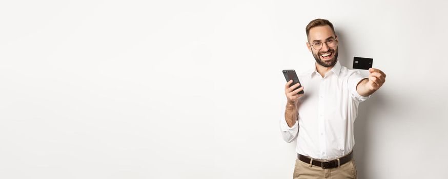 Business and online payment. Excited man showing his credit card while holding smartphone, standing satisfied against white background.