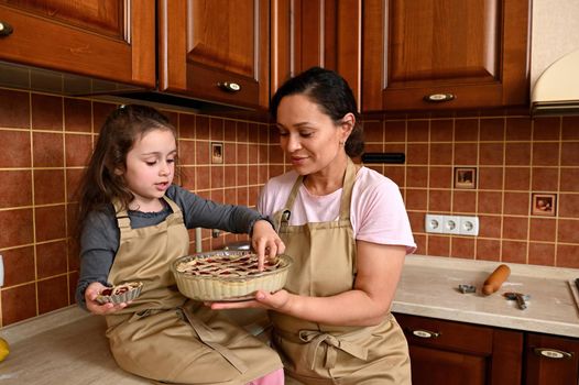 Adorable baby girl sitting on kitchen countertop, holding her first baked homemade cherry cake, enjoying cooking class with her loving mother. Pretty woman and her cute daughter cooking pie together