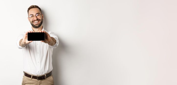 Happy business man showing mobile screen, holding phone horizontally, standing satisfied against white background.