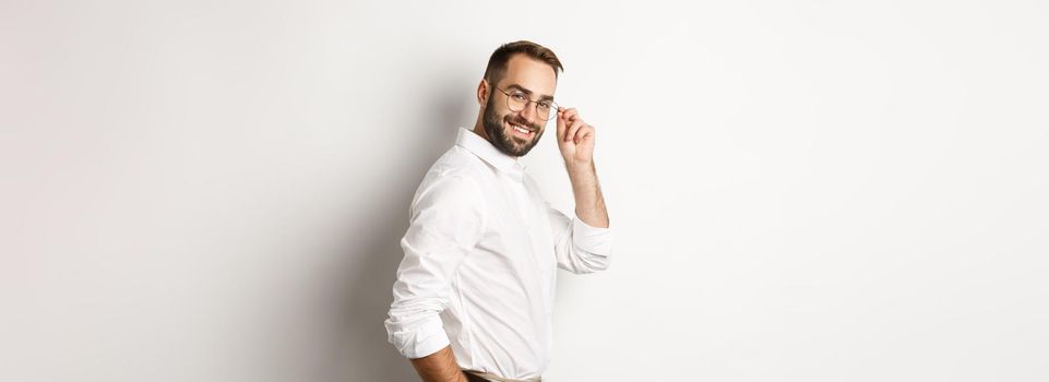 Handsome businessman turn at camera and looking confident, smiling cheeky, standing over white background.