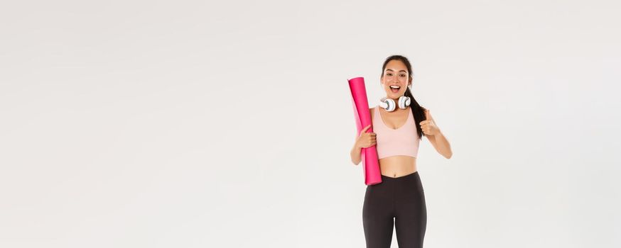 Full length of cheerful satisfied, slim asian girl with fitness rubber mat and headphones, female athlete showing thumbs-up, recommend gym or yoga classes, looking pleased after good workout.