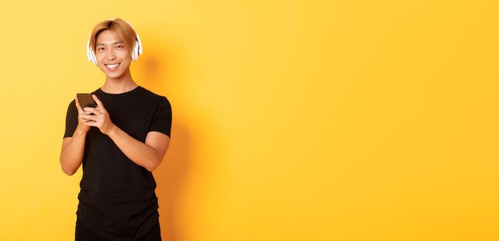 Good-looking smiling, pleased asian guy in wireless headphones, holding smartphone and looking satisfied camera, listening music or podcast.