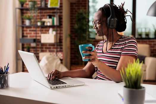 Smiling woman in wireless headphones watching movie online on laptop, holding mug, drinking coffee. Indoor entertainment, leisure activity on internet, teenager in earphones relaxing