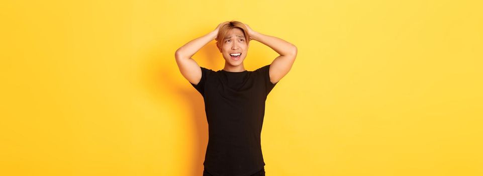 Portrait of young asian guy panicking, looking anxious and upset, holding hands on head and grimacing, standing over yellow background.