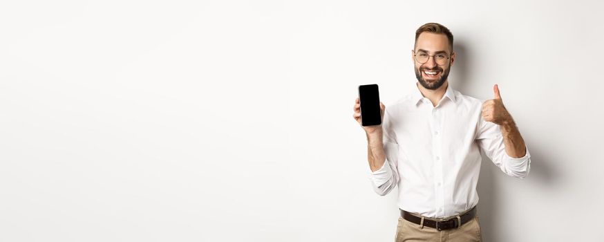 Satisfied business man in glasses showing thumbs up and demonstrating mobile phone screen, recommending app, standing over white background.