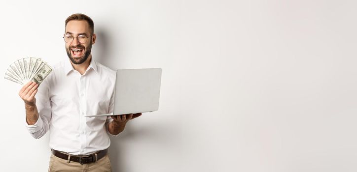 Business and e-commerce. Confident businessman showing how work online, winking, holding money and laptop, standing over white background.