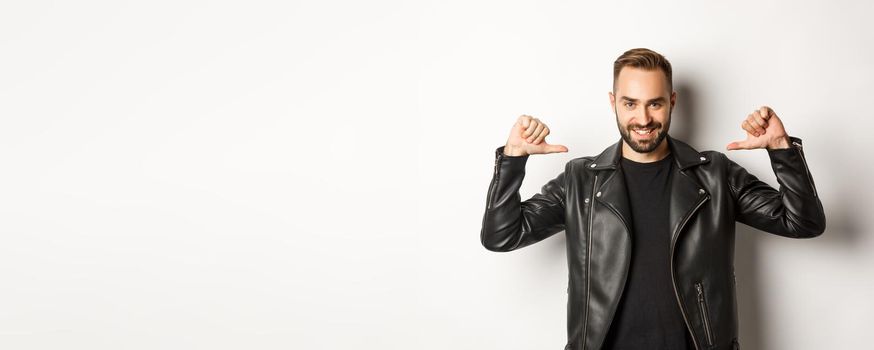 Confident handsome man wearing black leather jacket, pointing at himself and smiling self-assured, standing against white background.