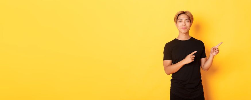 Smiling asian male student with blond hair, pointing fingers right, showing logo, standing yellow background.