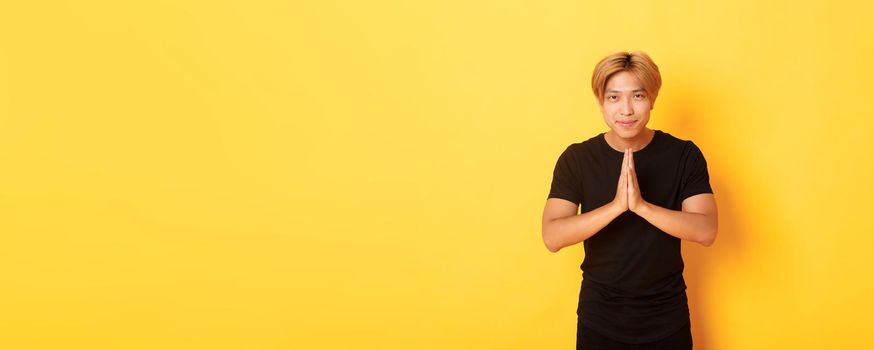 Portrait of friendly and polite asian blond guy, clasp hands together in namaste gesture, bowing and greeting person, standing over yellow background.