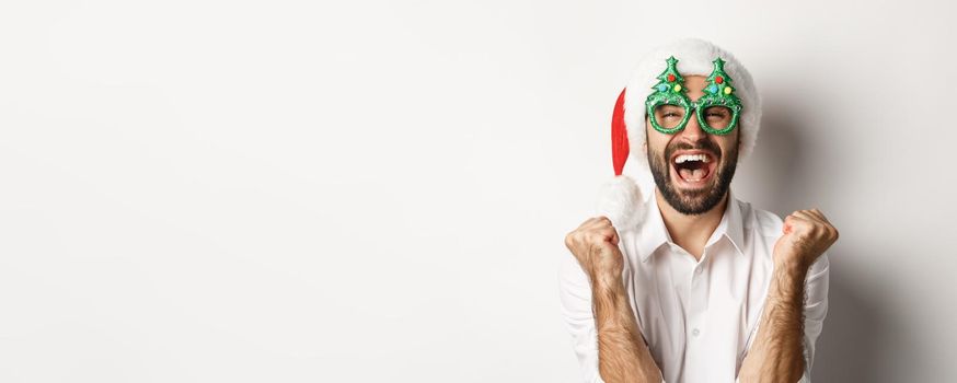 Close-up of man celebrating christmas or new year, wearing xmas party glasses and santa hat, rejoicing and shouting of joy, standing over white background.