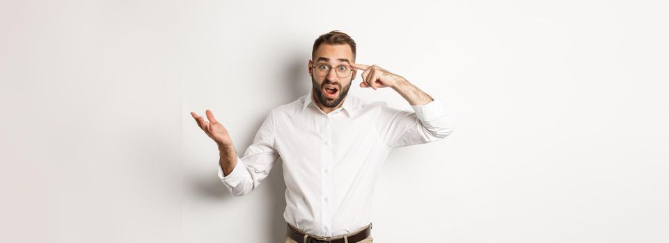 Confused and shocked man pointing at head, scolding employee for acting stupid, standing over white background.