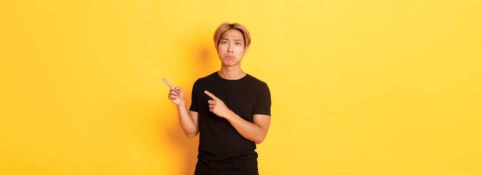 Portrait of disappointed and sad handsome blond asian guy, sulking upset, pointing fingers upper right corner, standing yellow background.