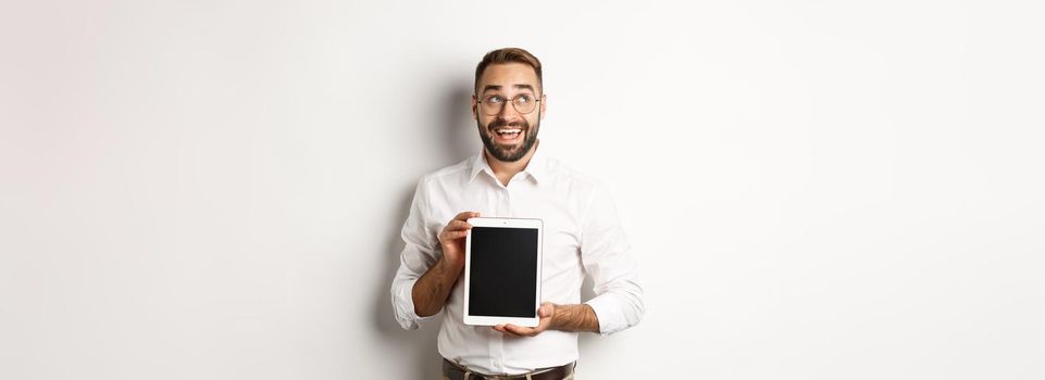 Shopping and technology. Thoughtful man showing digital tablet screen, looking at upper left corner and thinking, standing over white background.