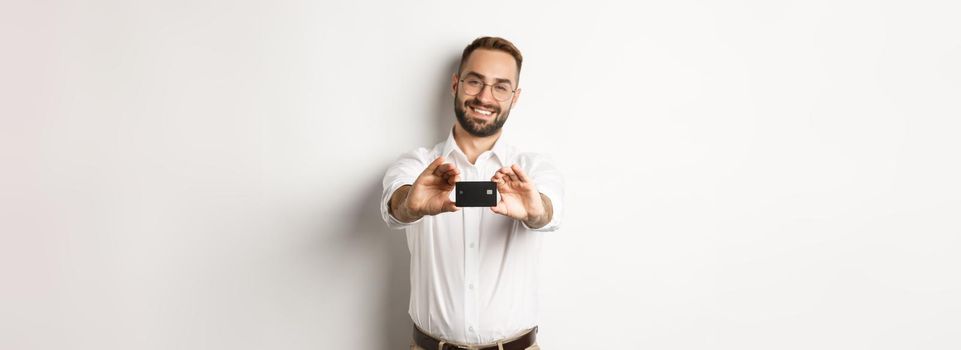 Handsome man in glasses holding a credit card, smiling pleased, standing over white background.