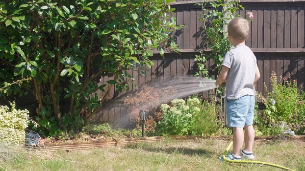 Funny little boy watering lawn plants in garden housing backyard. Adorable child playing with irrigation hose at hot sunny summer outdoors. Children help with housework. activity for kids. Childhood.